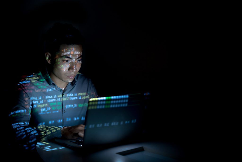 Man sits in dark room working on computer with code reflected on his face