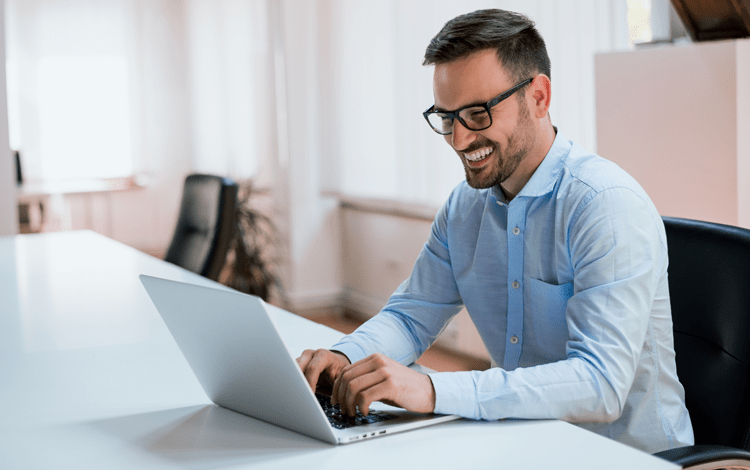 man with glasses, short beard and wearing a blue button down shirt smiles while typing on a laptop