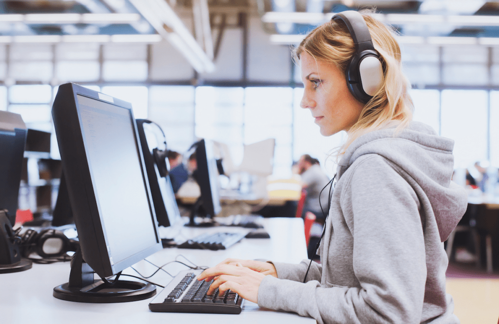 Woman with blonde hair, wearing grey hoodie and earphones works at a computer