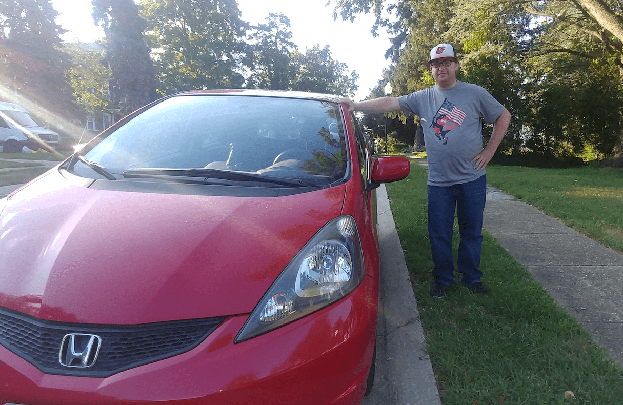 Man wearing Baltimore Orioles' shirt and hat leans against a red car