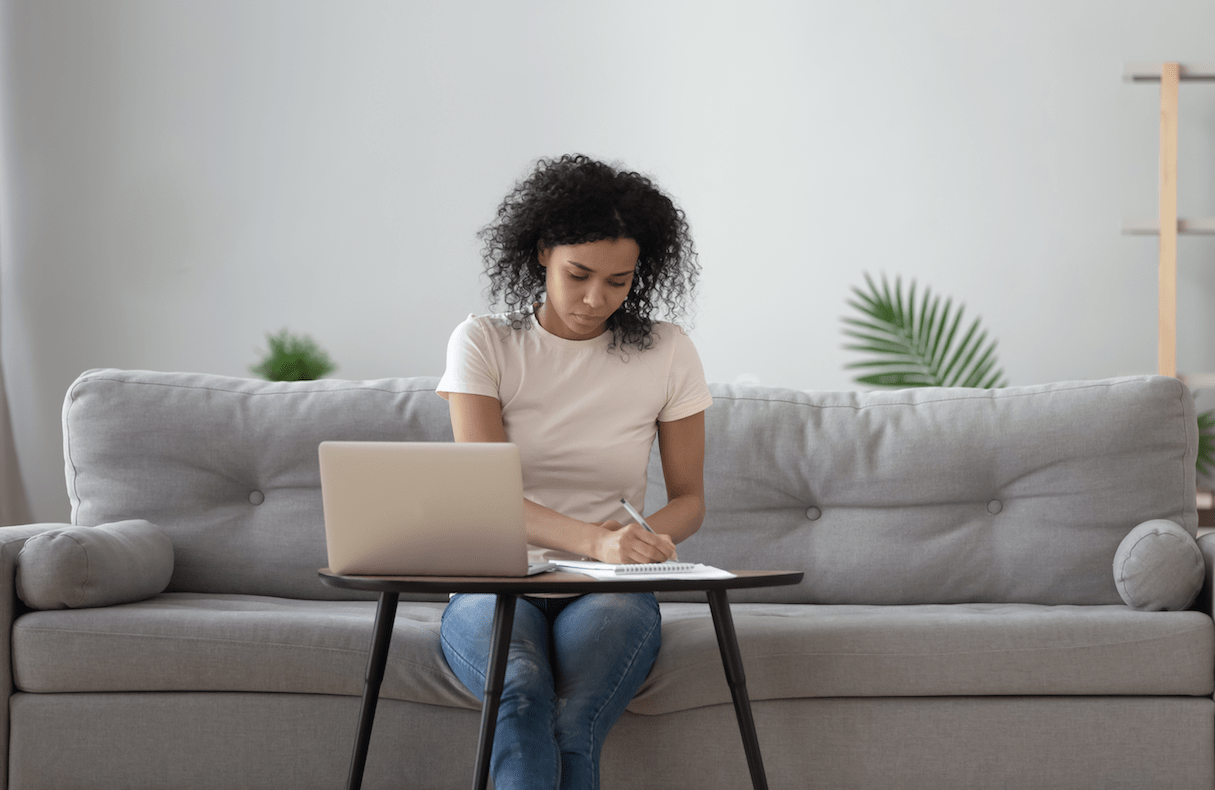Woman with curly hair sits on a grey couch writing in a notebook and working on a laptop computer