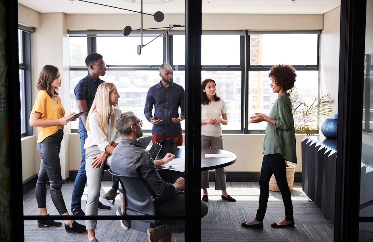 Woman standing in green shirt leads meeting of 6 people in conference room in high-rise office building