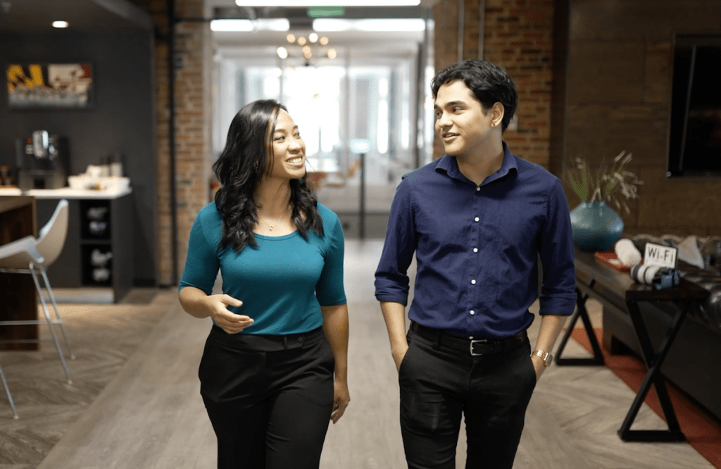 Woman with long dark hair, wearing a blue shirt, walks down an office hall with a man wearing blue button down shirt and black pants