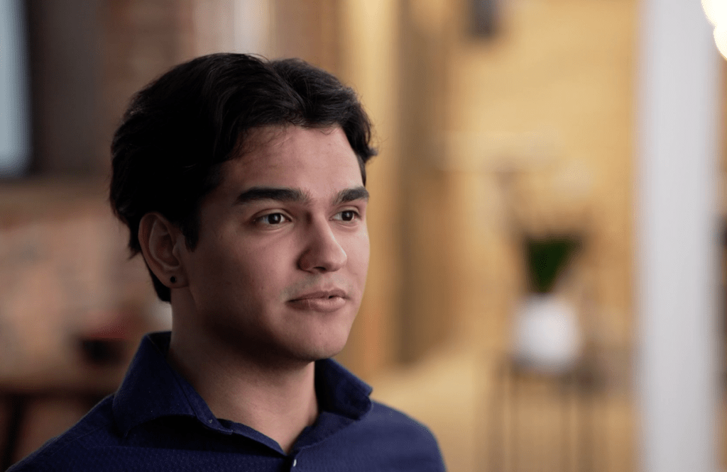 Man with blue shirt sits in chair for a video interview