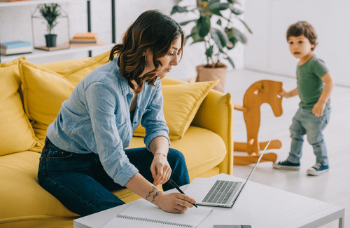 Woman trying to work from home on yellow couch as male child plays in the background