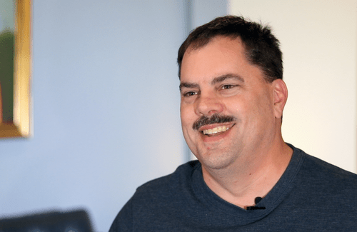 Man with mustache and blue shirt smiles at the camera during video interview