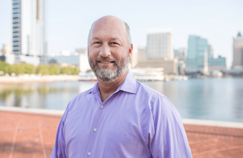 Man with grey beard and purple shirt smiles at the camera along Baltimore's Inner Harbor