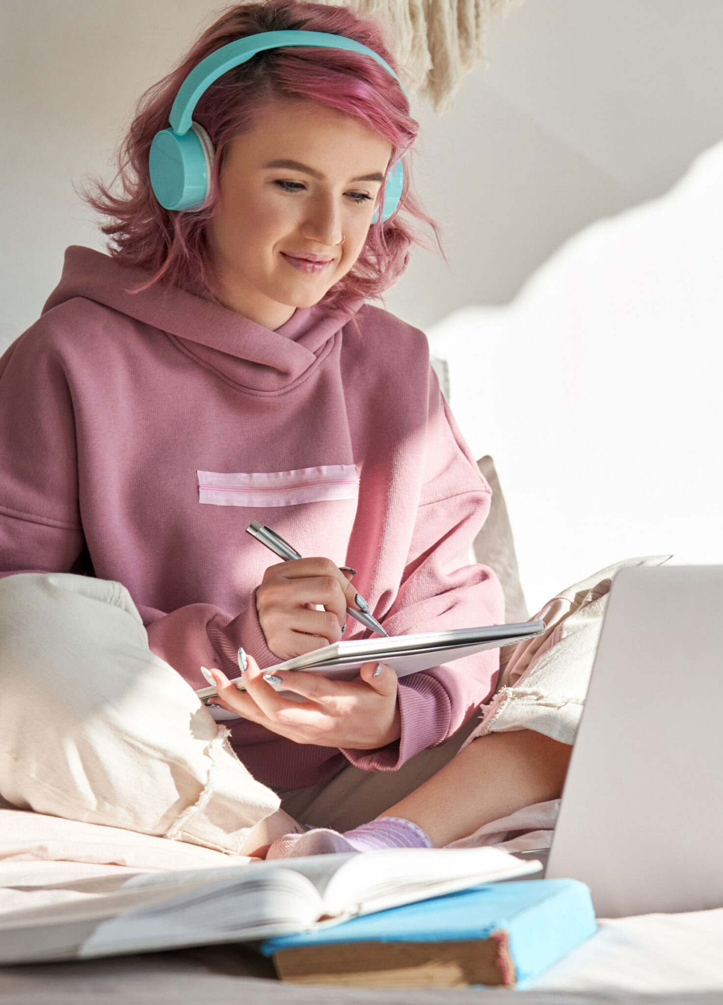 Teenage girl with pink hair and blue headphones sits on her bed working on computer,