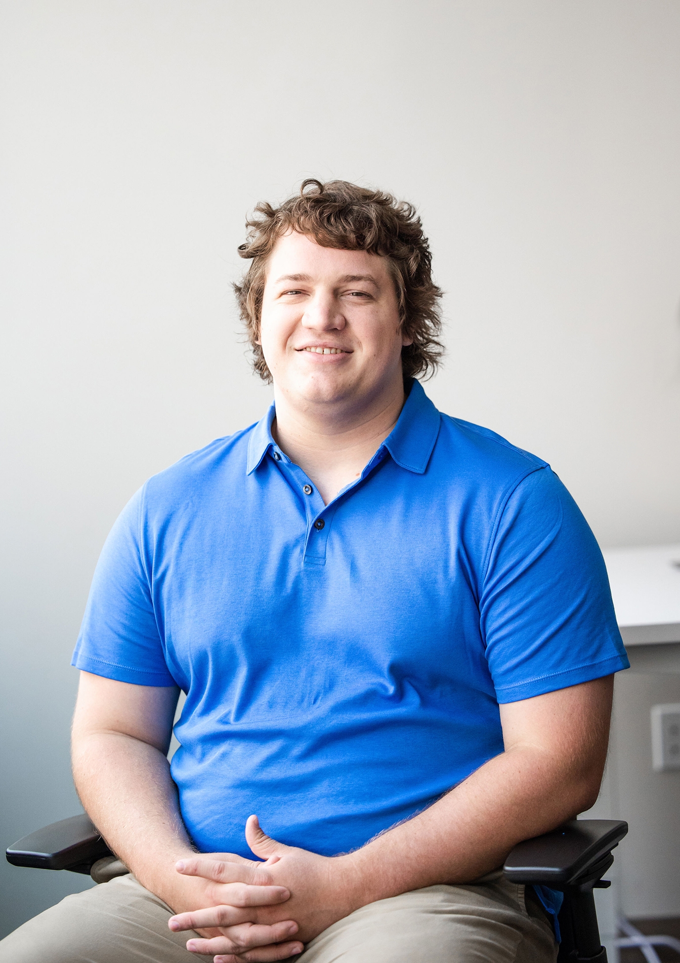 Man with medium length dark hair wearing a blue shirt sits in office chair smiling at camera