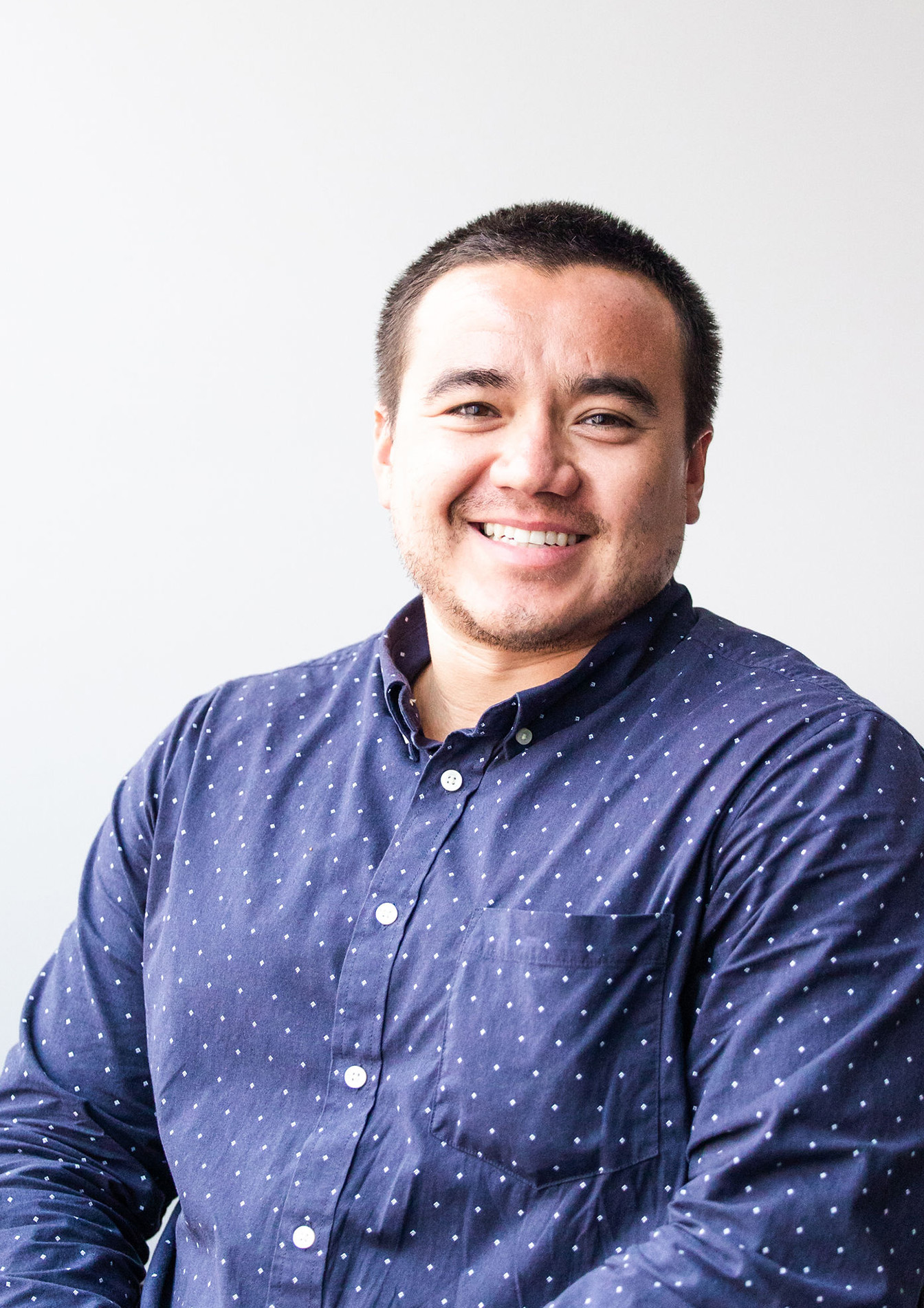 Man with short hair wearing blue shirt with white dots sits in office chair smiling at camera