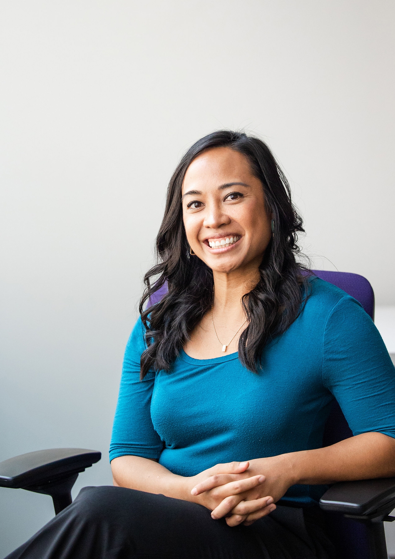 Woman with long, dark curly hair and a blue shirt sits smiling at the camera