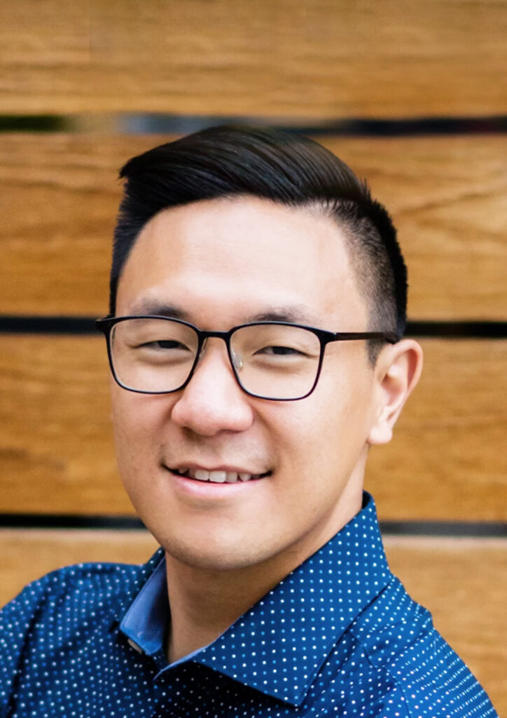 Man with short, black hair, glasses and white dotted blue shirt smiles looking at camera