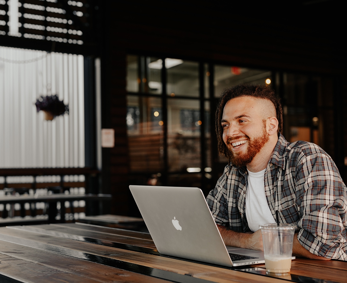 Man with locs and a flannel shirt works on an Apple laptop from a coffee shop