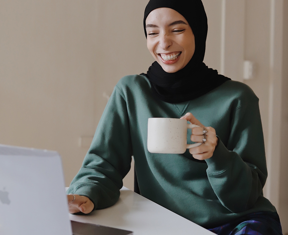 Woman hearing a hijab and holding a mug works on a laptop computer