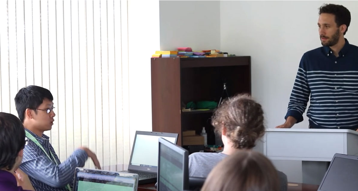 Man with beard stands at lecture in front of a class for software developers