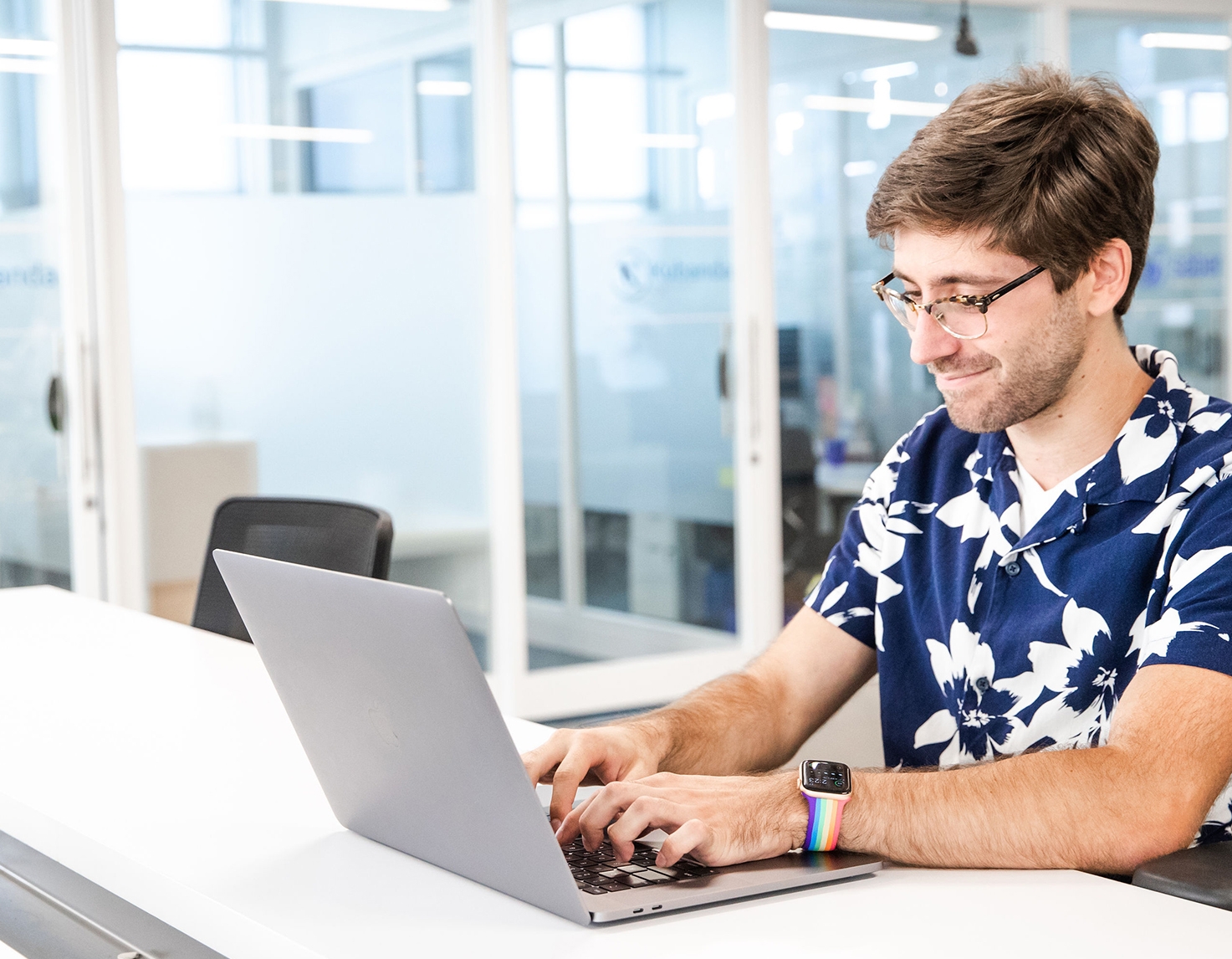 Man with short brown hair, wearing a blue floral pattern shirt works on a laptop in a bright office