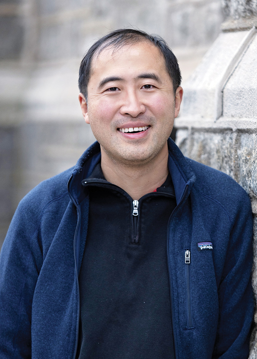 Man with short black hair wearing a blue zipper fleece leans against a stone wall smiling at the camera