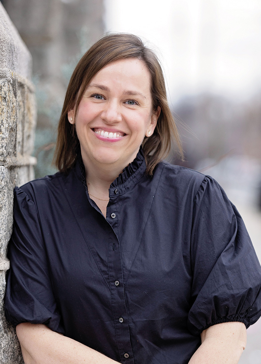 Woman with medium-length brown hair and dark blue shirt leans against a stone wall smiling at the camera