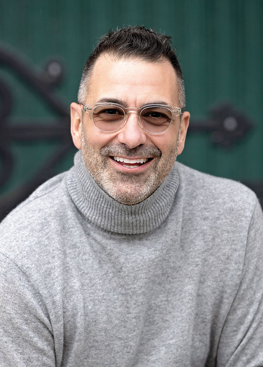 Man with short hair, glasses and wearing a grey turtle neck sweater smiles at the camera