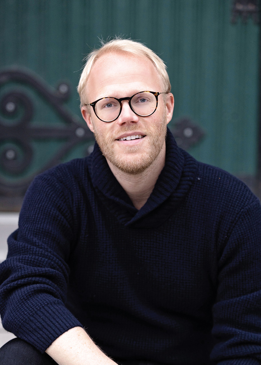 Man with short blonde hair and glasses, wearing dark blue sweater sits and smiles at camera