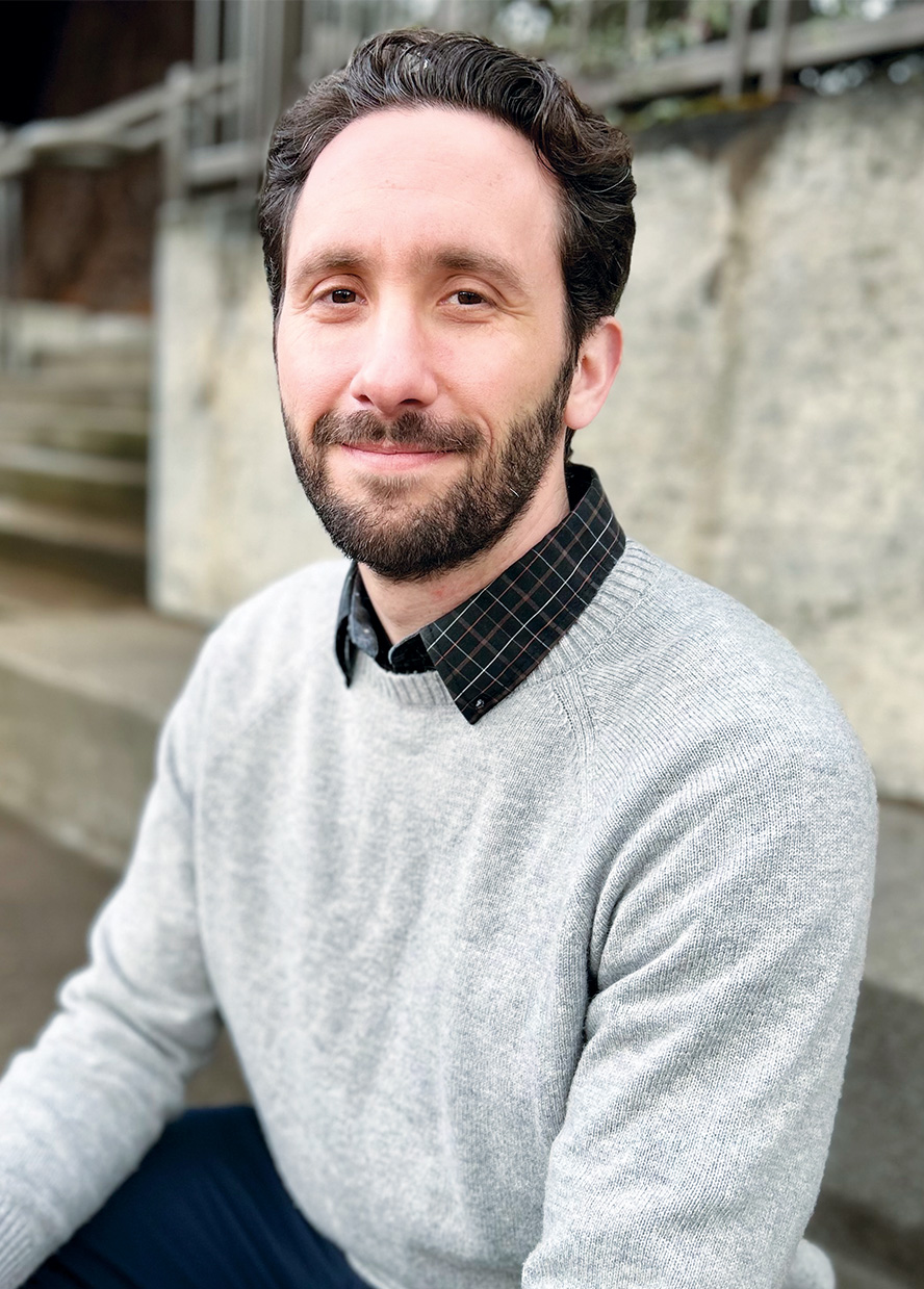 Man with short wavy hair and beard, wearing a grey sweater smiles at the camera