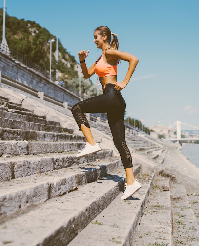 Woman in black workout leggings and a ponytail runs up steps in an outdoor stadium.