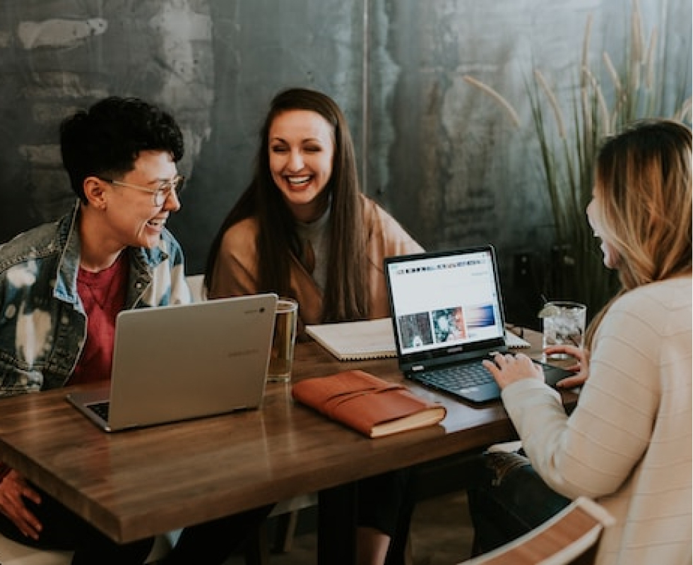 Three people sit at a restaurant table working on laptops and laughing with each other