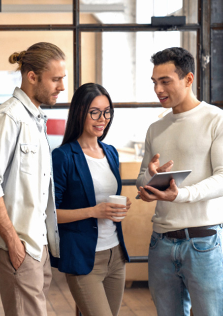 Man with manbun, women with glasses holding coffee mug look at tablet held by man with long-sleeve white shirt