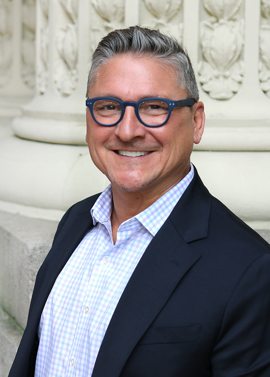 Man with salt-and-pepper hair, wearing glasses and a dark blue sport coat smiles at the camera in front of a Grecian column