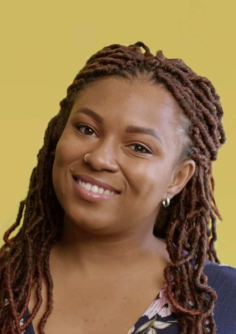 Woman with locs and a blue shirts smiles with yellow background