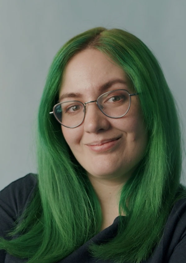 Woman with green hair, glasses and blue shirt smiles at camera against a blue background