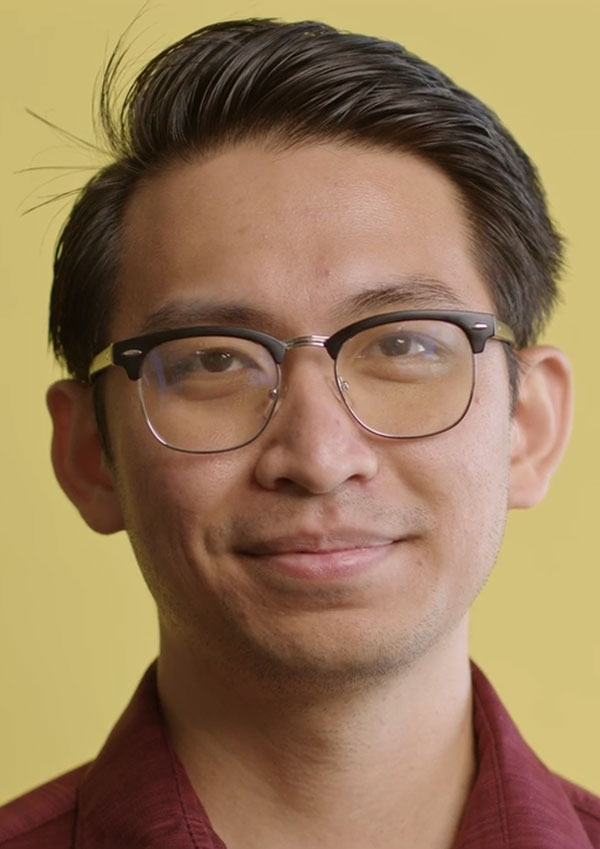 Man with short, dark hair wearing glasses and a red collared shirt stands in front of a yellow backdrop