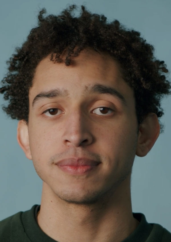 Man with short, dark curly hair looks at camera against a blue background