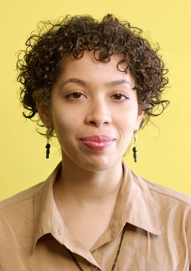 Woman with short, dark curly hair smiles at the camera against a yellow background