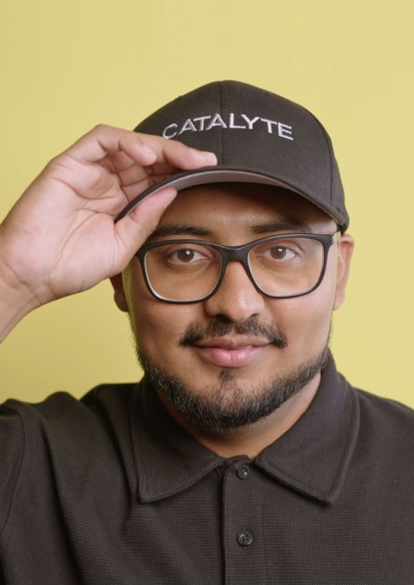 Man with glasses, beard and grey polo shirt touched the bill on his baseball cap while looking at the camera against a yellow