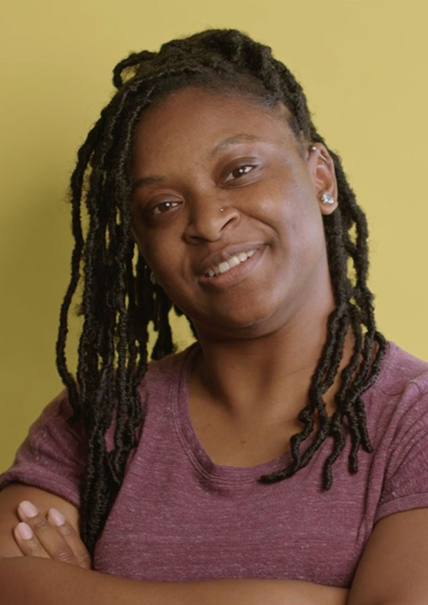 Woman with locs and purple shirt stands with arms crossed looking at camera against a yellow background