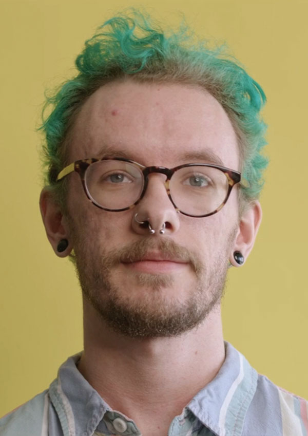 Man with short green hair, glasses, beard and piercing looks at camera against a yellow background
