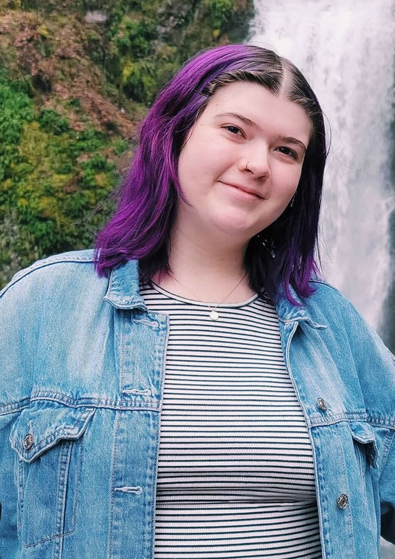 Woman with purple hair and a nose ring, wearing striped shirt and jean jacket with waterfall in the background