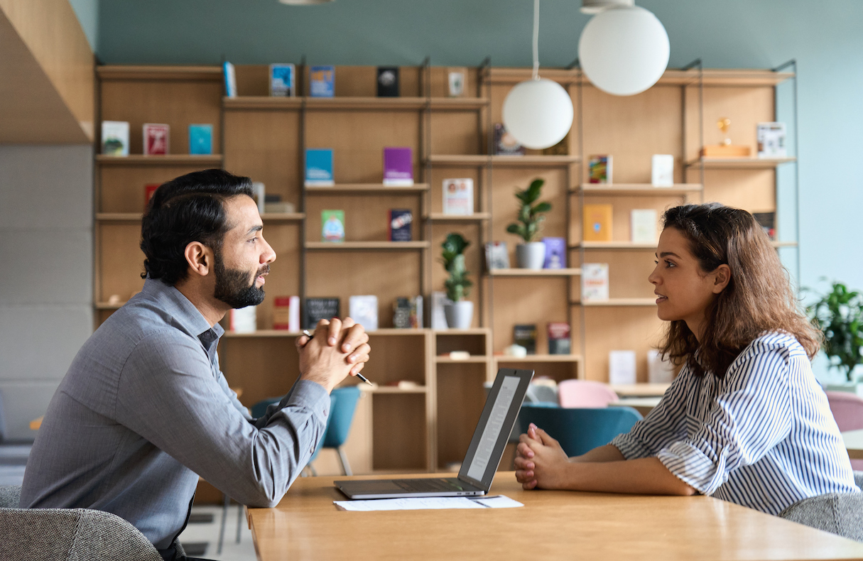 Man with dark hair and a beard interviewing female job candidate with brown hair and a striped shirt across a desk