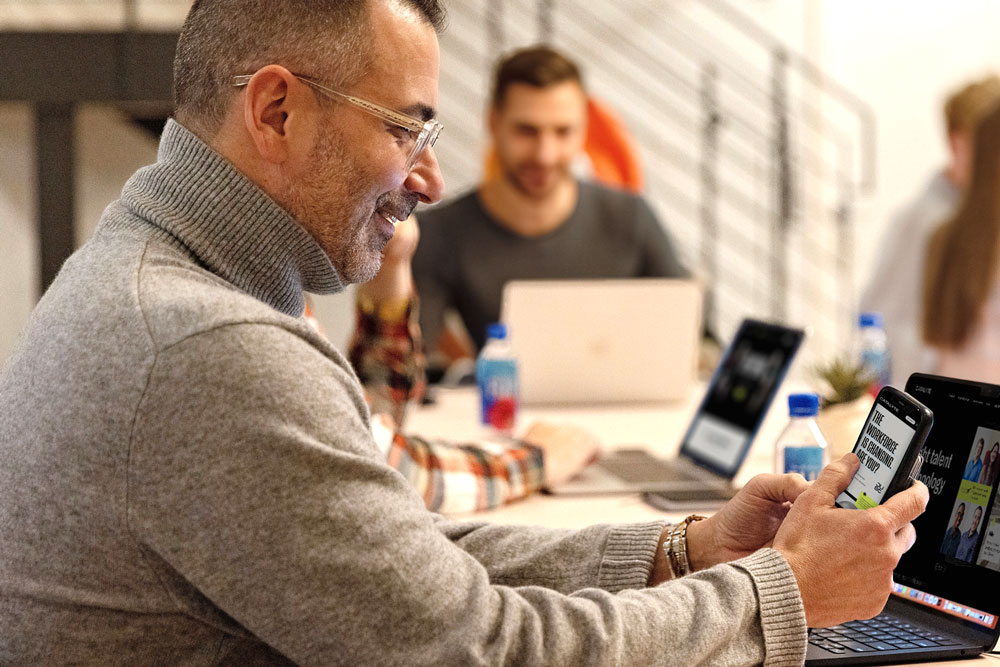 Man in a gray shirt with glasses sits at a conference table looking at a website on his phone