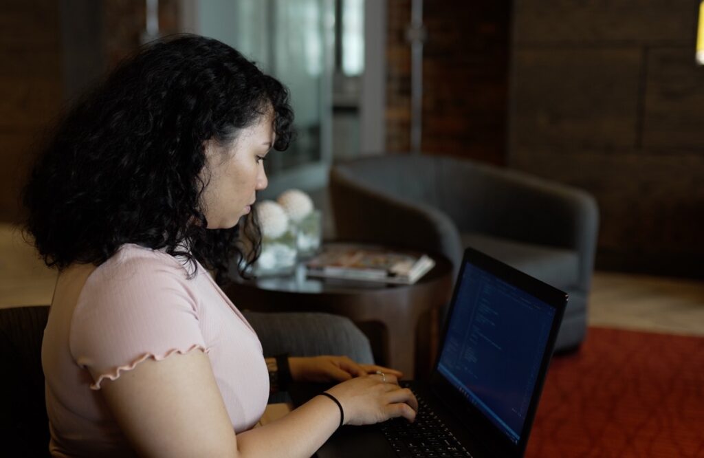 Woman with long dark curly hair and a pink top sits in an open office working on a laptop computer